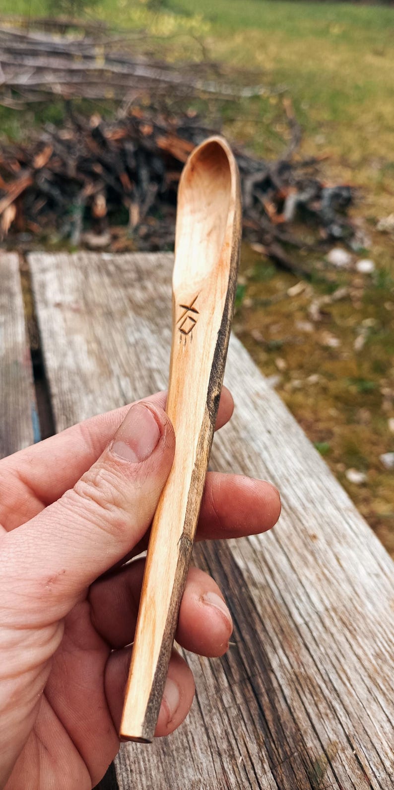 K&ouml;nnte beinhalten: Handgeschnitzter Holzl&ouml;ffel mit nat&uuml;rlicher, rustikaler Optik. Der L&ouml;ffel hat einen langen Stiel und eine abgerundete Sch&uuml;ssel mit einem strukturierten Rindenrand. Ein kleines Symbol ist in den Griff geschnitzt. Der L&ouml;ffel wird vor einem verschwommenen Hintergrund im Freien gehalten.