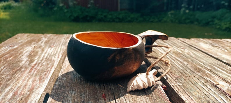 May include: A handcrafted wooden kuksa cup featuring a dark, charred exterior finish and a natural, light-colored interior bowl. The cup includes a carved handle with a small, light-colored wooden leaf-shaped charm attached by a thin twine cord.