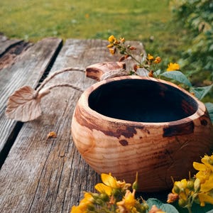 May include: A round wooden cup with a dark interior and natural grain patterns on the exterior. The cup features a carved handle with a jute cord attached to a small wooden leaf-shaped charm, showcasing a rustic and handcrafted aesthetic.
