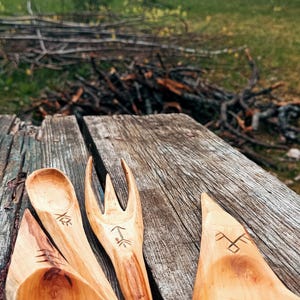 K&ouml;nnte beinhalten: Handgeschnitztes Holzgeschirr, darunter ein L&ouml;ffel, eine Gabel und ein Spatel, mit ge&auml;tzten Designs. Das hellbraune Geschirr ist auf einer verwitterten Holzoberfl&auml;che angeordnet. Im Hintergrund befindet sich eine rustikale rote Scheune.