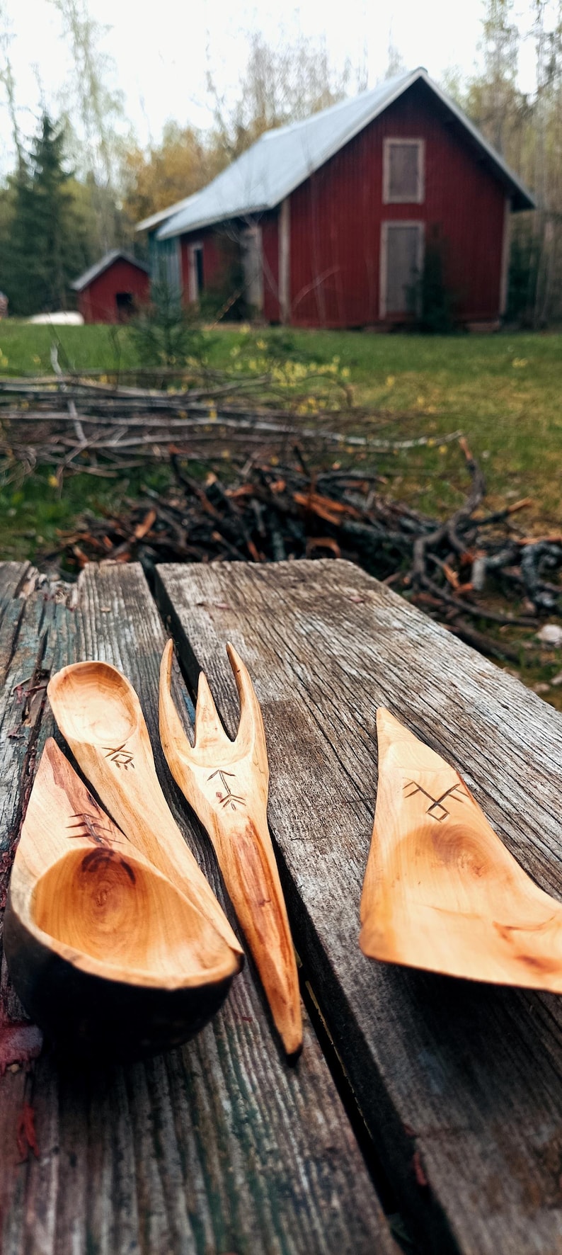 K&ouml;nnte beinhalten: Handgeschnitztes Holzgeschirr, darunter ein L&ouml;ffel, eine Gabel und ein Spatel, mit ge&auml;tzten Designs. Das hellbraune Geschirr ist auf einer verwitterten Holzoberfl&auml;che angeordnet. Im Hintergrund befindet sich eine rustikale rote Scheune.