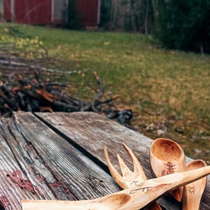 K&ouml;nnte beinhalten: Handgeschnitztes K&uuml;chenutensilien-Set aus Holz, darunter eine Gabel, ein L&ouml;ffel und ein Servierl&ouml;ffel, liegen auf einer verwitterten Holzoberfl&auml;che. Im Hintergrund ist eine rote Scheune zu sehen. Die Holzmaserung und die Schnitzereien sind sichtbar.