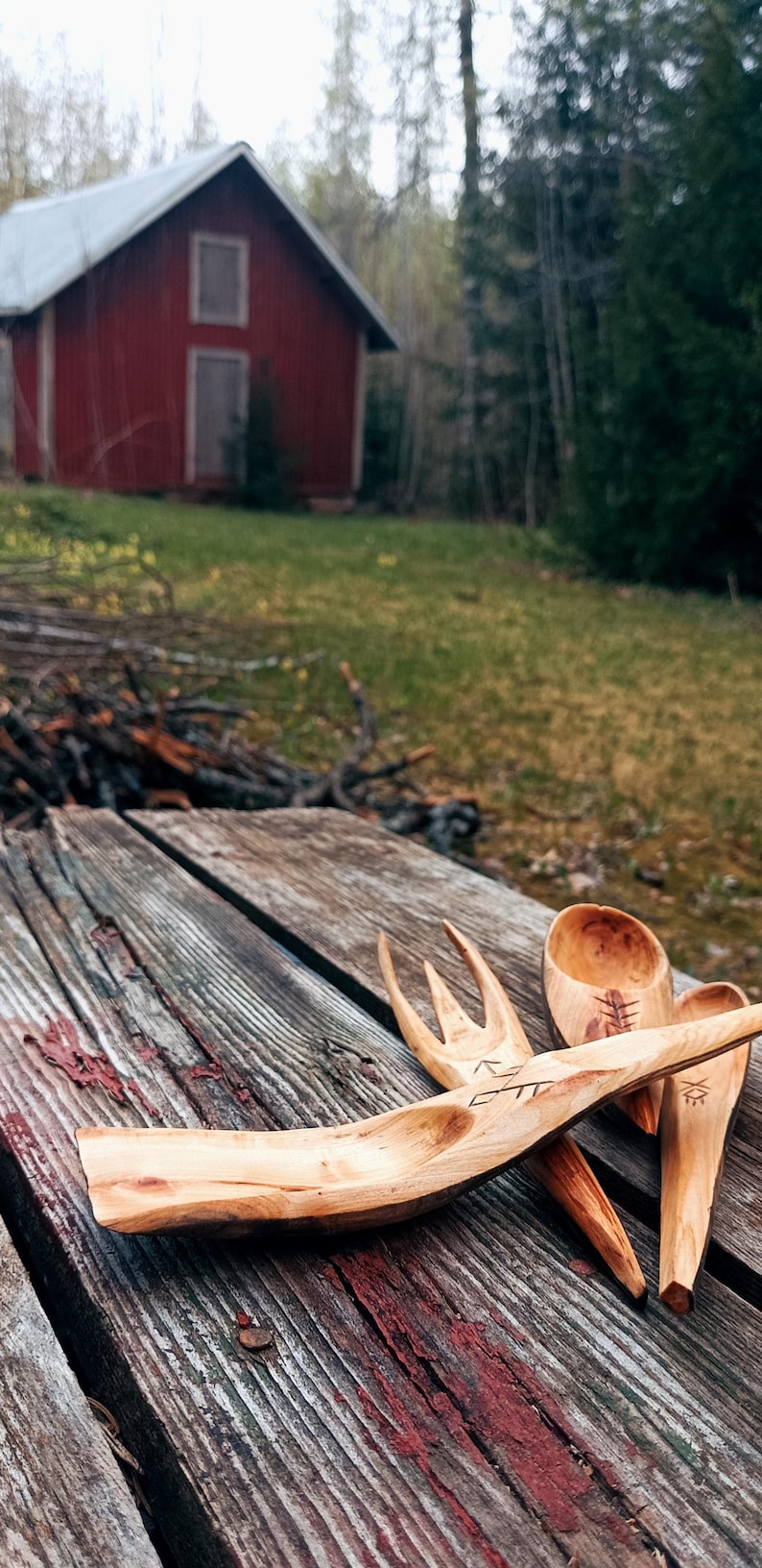 K&ouml;nnte beinhalten: Handgeschnitztes K&uuml;chenutensilien-Set aus Holz, darunter eine Gabel, ein L&ouml;ffel und ein Servierl&ouml;ffel, liegen auf einer verwitterten Holzoberfl&auml;che. Im Hintergrund ist eine rote Scheune zu sehen. Die Holzmaserung und die Schnitzereien sind sichtbar.