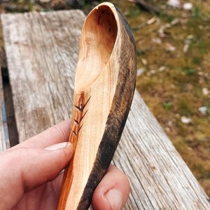 K&ouml;nnte beinhalten: Handgeschnitzter Holzl&ouml;ffel mit nat&uuml;rlichem Finish. Der L&ouml;ffel hat ein geschnitztes Design am Griff und einen dunklen, kontrastierenden Rand. Der L&ouml;ffel wird vor einer verwitterten Holzoberfl&auml;che gehalten.