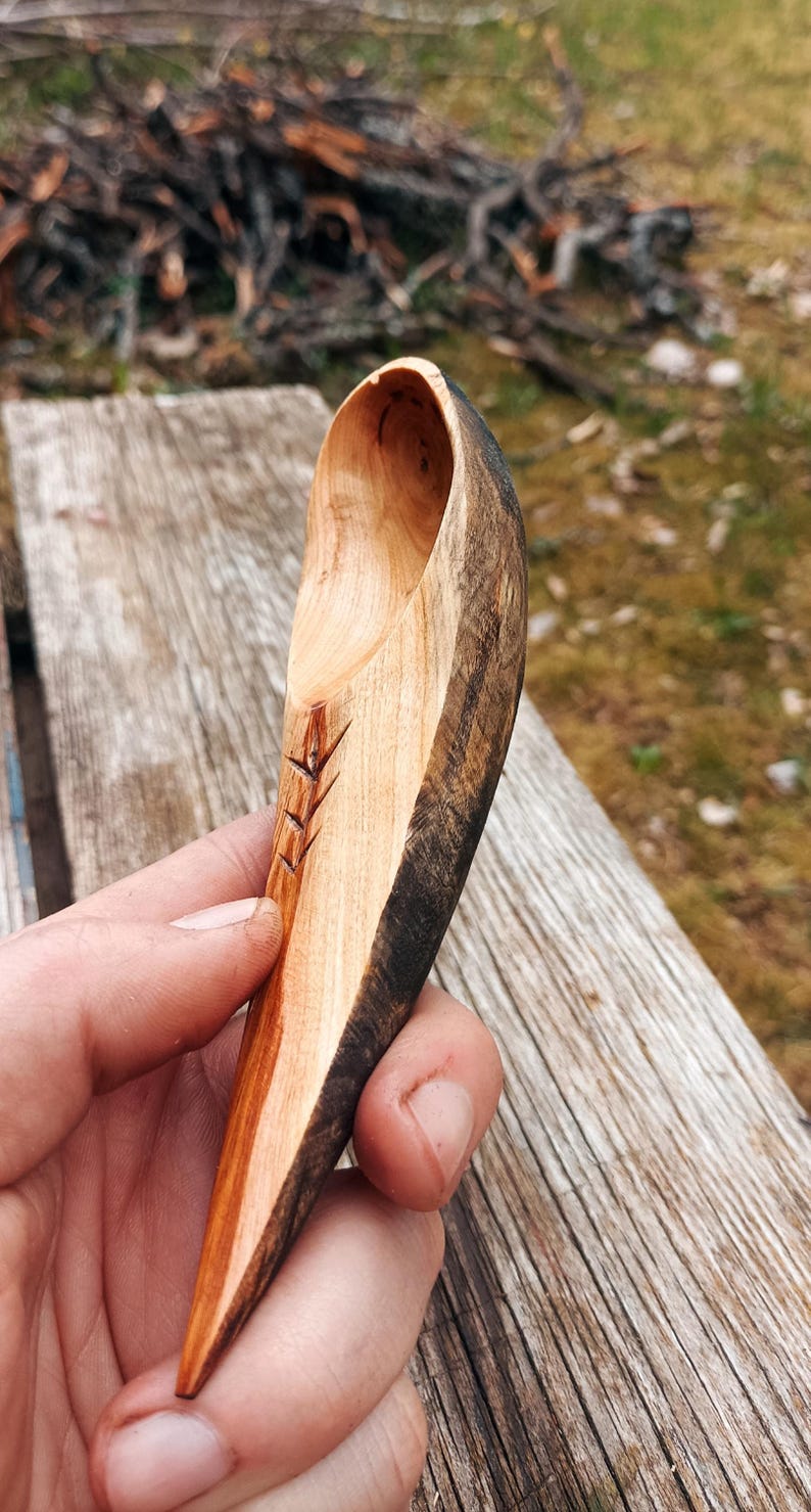 K&ouml;nnte beinhalten: Handgeschnitzter Holzl&ouml;ffel mit nat&uuml;rlichem Finish. Der L&ouml;ffel hat ein geschnitztes Design am Griff und einen dunklen, kontrastierenden Rand. Der L&ouml;ffel wird vor einer verwitterten Holzoberfl&auml;che gehalten.