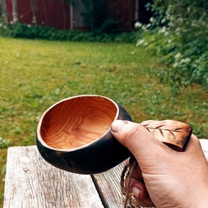 May include: A hand-carved wooden kuksa cup with a dark exterior and a natural light brown interior, featuring a handle with a carved leaf pattern and a matching wooden spoon attached by a thin twine cord with a small wooden leaf charm.