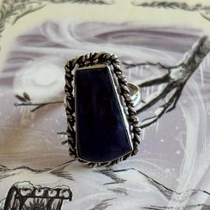 May include: A silver ring featuring a large, rectangular, dark blue stone. The stone is set within a twisted rope-like border. The ring has a simple band and is displayed against a winter-themed background.
