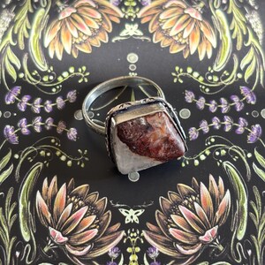 Puede incluir: Un anillo de plata con una piedra rectangular de color marrón rojizo engastada en un bisel cuadrado de plata. El anillo descansa sobre una superficie negra decorada con ilustraciones florales y botánicas en tonos naranja, rosa, verde y morado.