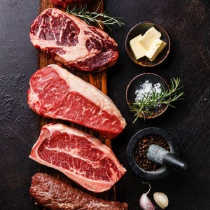 Könnte beinhalten: Draufsicht auf verschiedene rohe Rindfleischstücke auf einem Holzbrett. Dazu Butter, Salz, Pfefferkörner, Rosmarinzweige und Knoblauchzehen. Das Bild zeigt verschiedene Steak-Cuts. Der Hintergrund ist dunkel.