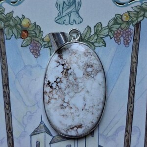 Może przedstawiać: Owalny wisiorek z białym kamieniem z brązowymi żyłkami, osadzony w srebrnej metalowej ramie. Wisiorek wisi na srebrnej pętli. Tło zawiera akwarelową ilustrację z winogronami i budynkiem.