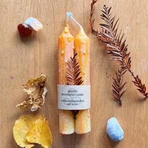 Op de afbeelding: Twee oranje en witte kaarsen met een label met de tekst "apollo devotional candle celticcrosscandles est 2024".