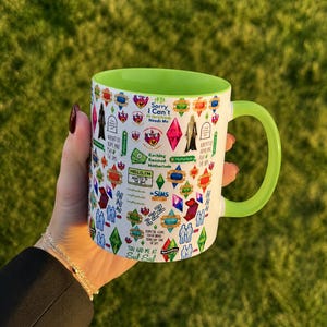 May include: A white ceramic mug with a bright lime green interior and handle, featuring a colourful collage of The Sims video game icons, plumbobs, and text including Sorry I Can't My Sims Need Me, Kaching, Rosebud, Motherlode, and You Had Me At Sul Sul.