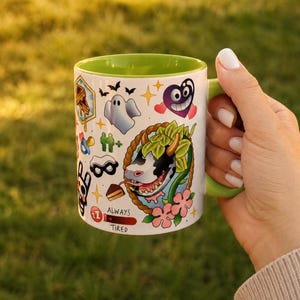 Pode incluir: Uma caneca de cer&acirc;mica branca com interior e al&ccedil;a verde-lim&atilde;o, apresentando uma estampa gr&aacute;fica colorida de uma cabe&ccedil;a de vaca, um fantasma, um cora&ccedil;&atilde;o roxo e v&aacute;rios &iacute;cones pequenos, incluindo uma fatia de bolo e o texto ALWAYS TIRED.