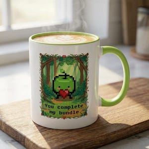 Könnte beinhalten: Weiße Keramik-Tasse mit grünem Henkel und Rand, mit einem pixeligen grünen Apfel, der ein rotes Herz hält. Die Tasse trägt den Text "You complete my bundle" und ist mit einem Latte gefüllt.
