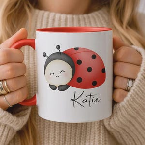 Peut inclure: Mug en céramique blanche avec intérieur et anse rouges. Le mug présente un dessin de coccinelle de dessin animé avec un visage souriant et le nom "Katie" en écriture noire. La coccinelle a une carapace rouge à pois noirs.