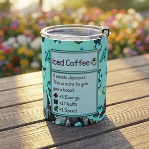 Puede incluir: Un vaso t&eacute;rmico turquesa con tapa negra. El vaso presenta gr&aacute;ficos pixelados y el texto "Iced Coffee". El texto contin&uacute;a: "It smells delicious. This is sure to give you a boost. +3 Energy, +1 Health, +1 Speed."