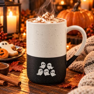 May include: A two-toned ceramic mug with a speckled cream top and a solid black bottom. The black section features five small white ghost illustrations. The mug is filled with a hot beverage topped with whipped cream and a dusting of cinnamon.