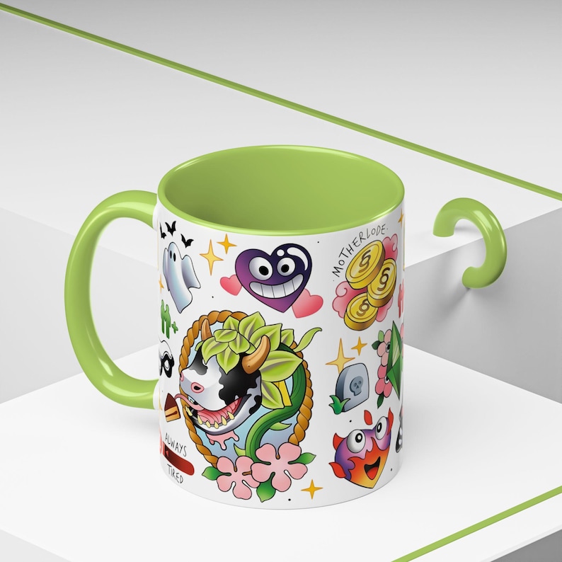 Pode incluir: Uma caneca de cer&acirc;mica branca com interior e pega em verde-lima, apresentando uma impress&atilde;o gr&aacute;fica colorida de uma vaca, um cora&ccedil;&atilde;o roxo, moedas de ouro com a etiqueta MOTHERLODE, um fantasma e outros &iacute;cones. O texto ALWAYS TIRED est&aacute; em vermelho.
