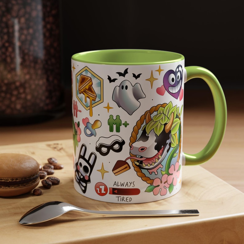May include: White ceramic mug with a green interior and handle, decorated with colorful tattoo-style illustrations. Designs include a ghost, heart, and a cow with a floral border. The mug also has the text "ALWAYS TIRED".