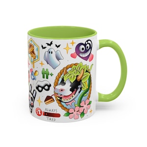 Pode incluir: Uma caneca de cer&acirc;mica branca com interior e al&ccedil;a verde-lim&atilde;o, apresentando uma colagem colorida em estilo tatuagem de uma vaca, um fantasma, um cora&ccedil;&atilde;o roxo, um waffle e outros &iacute;cones, incluindo uma barra de progresso vermelha com o texto ALWAYS TIRED.