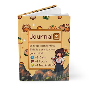 Peut inclure: Un journal à couverture rigide avec un design pixélisé représentant une femme tenant une tasse, des citrouilles et des crabes. La couverture du journal porte le mot "Journal" et le texte "It feels comforting. This is sure to clear your mind."
