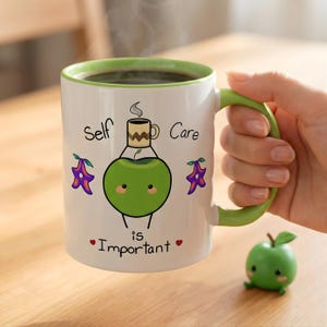 May include: White ceramic mug with a green interior and handle, featuring the text "Self Care is Important." The mug has a cartoon apple design with a coffee cup on top and purple star-shaped accents. A small green apple figurine is next to the mug.
