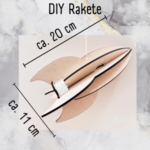 Może przedstawiać: DIY Rakete, zestaw do samodzielnego montażu rakiety z drewna, ma około 11 cali wysokości i 20 cali szerokości. Rakieta wykonana jest z jasnobrązowego drewna i ma biały papierowy tubus w środku.