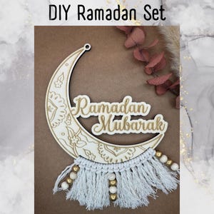 Könnte beinhalten: Ein Holz-Halbmond mit den Worten "Ramadan Mubarak" in goldenen Lettern. Der Mond ist mit einem weißen und goldenen, filigranen Design verziert. Ein weißer Makramee-Quaste mit goldenen Perlen hängt am unteren Ende des Mondes.