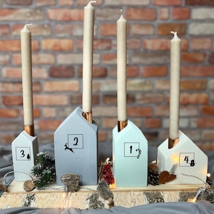 Op de afbeelding: Adventskalender met vier huisvormige kaarshouders in blauwe, grijze en witte tinten. Elke houder heeft een koperkleurige kaarsenbasis en een lange, crèmekleurige kaars. De huizen zijn genummerd van 1 tot en met 4.