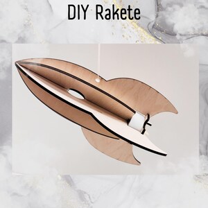 Może przedstawiać: Drewniany zestaw do samodzielnego wykonania rakiety z napisem "DIY Rakete" na g&oacute;rze. Rakieta wykonana jest z jasnobrązowego drewna i ma białą papierową rurkę z tyłu.