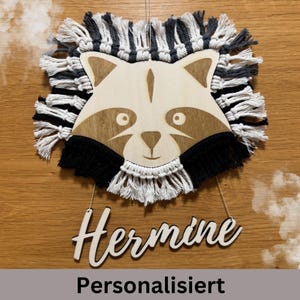 Può includere: Decorazione da parete a forma di procione in legno con frangia in macramè nera, bianca e grigia. Il viso del procione è marrone chiaro con dettagli marrone scuro. Il nome "Hermine" è visualizzato sotto. La parola "Personalizzato" è in basso.