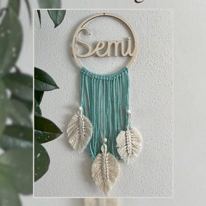 Può includere: Un acchiappasogni in macramè turchese con un anello di legno su cui è scritto "Semi" in lettere bianche. L'acchiappasogni ha tre piume in macramè bianche che pendono da esso.