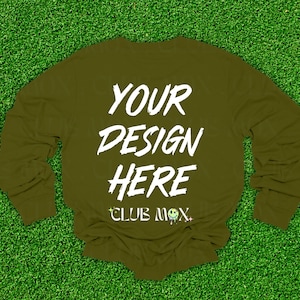 May include: Olive green long-sleeve shirt with the text "YOUR DESIGN HERE" in white, along with "CLUB MOX" and a small alien graphic. The shirt is displayed on a green turf background.