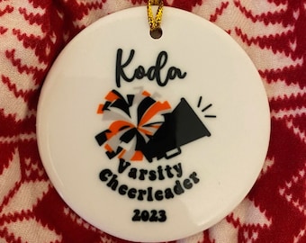 May include: White ceramic ornament with the name "Koda" written in black cursive. It features a black and orange cheerleader pom-pom and a black megaphone with a white star. The text "Varsity Cheerleader 2023" is written in black below the pom-pom and megaphone.