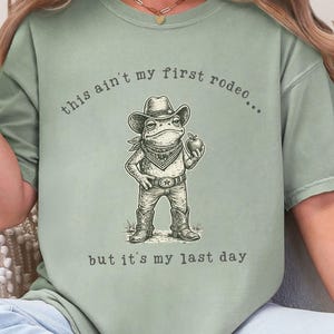 May include: Sage green t-shirt featuring a cartoon frog dressed as a cowboy, holding an apple. The text on the shirt reads "this ain't my first rodeo... but it's my last day."