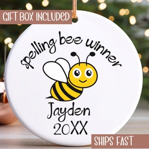 May include: A white ceramic ornament with a cartoon bee design. The text "spelling bee winner" arches above the bee, with the name "Jayden" and the year "20XX" below. The ornament includes a gold string for hanging.