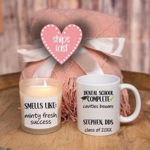 May include: A gift set featuring a white mug with "DENTAL SCHOOL COMPLETE" text, a candle labeled "SMELLS LIKE: minty fresh success", and a pink blanket tied with a white ribbon. The mug includes the name "STEPHEN, DDS" and class year.