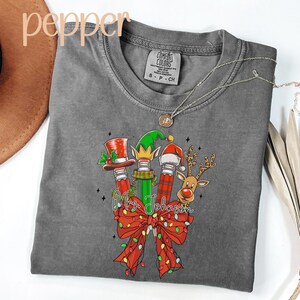 May include: A gray Comfort Colors t-shirt featuring a Christmas-themed graphic. The design includes pencils adorned with a top hat, elf hat, and Santa hat, along with a reindeer. The text "Mrs. Johnson" is written across a red bow with string lights.