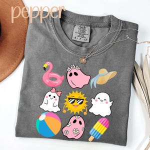 May include: A gray Comfort Colors t-shirt featuring summer-themed graphics. The design includes a pink flamingo float, ghosts, a sun with sunglasses, a beach ball, a sun hat, and an ice cream bar. A silver necklace is draped across the shirt.