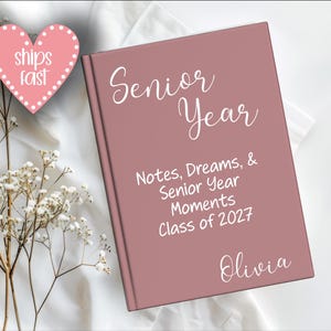 May include: A dusty rose-colored notebook with the words "Senior Year" in white script. The notebook also includes the text "Notes, Dreams, & Senior Year Moments Class of 2027 Olivia." A pink heart-shaped sticker with the words "ships fast" is also present.