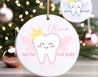 May include: White ceramic Christmas ornament with a cartoon tooth design. The tooth has a yellow crown and pink wings. Text reads "Olivia lost her first tooth!" and "20??". A smaller ornament reads "Oliver lost his first tooth!" and "2025".