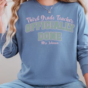 May include: A long-sleeved, blue shirt with the text "Third Grade Teacher OFFICIALLY DONE Mrs. Johnson" in pink and green. The shirt is a solid blue color and has a crew neck.