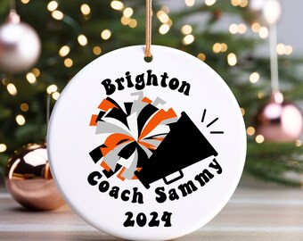 May include: White ceramic ornament with a gold string for hanging. The ornament features the text "Brighton Coach Sammy 2024" in black, along with a cheerleader megaphone and pom-poms in black, orange, and gray.