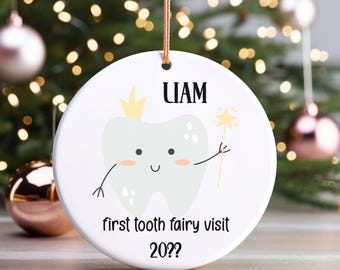 May include: A white ceramic Christmas ornament with a tooth fairy theme. The ornament features a cartoon tooth with a crown and a wand, the name "LIAM", and the text "first tooth fairy visit 20??". A gold string is attached for hanging.