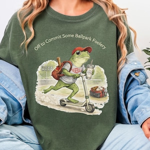 May include: Green t-shirt with the text "Off to Commit Some Ballpark Foolery." The shirt features a cartoon frog wearing a baseball cap and shirt, riding a scooter. The frog carries a drink and a backpack with a baseball bat.