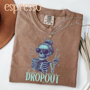 May include: Espresso-colored t-shirt featuring a skeleton wearing sunglasses and holding a drink, with the text "Bushover Teacher Dropout". The shirt has a Comfort Colors tag and a silver necklace.