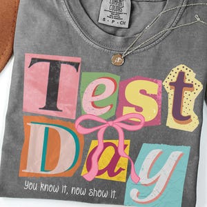 May include: A gray t-shirt with the words "Test Day" in colorful, block letter cut-out style. A pink bow is centered below the words. The text "You know it, now show it" is below the words. A silver necklace with a small pendant is on the shirt.