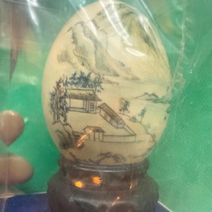 May include: An egg-shaped decorative object with a landscape scene painted on its surface. The artwork depicts mountains, trees, and buildings in a minimalist style. The egg sits on a dark brown wooden stand, showcasing the intricate details of the painted design.