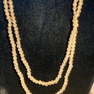 Puede incluir: Un collar largo de doble hebra con pequeñas cuentas redondas y translúcidas de color melocotón pálido. Las cuentas son de tamaño y forma uniformes, ensartadas para crear un bucle continuo y delicado que cuelga en dos capas.