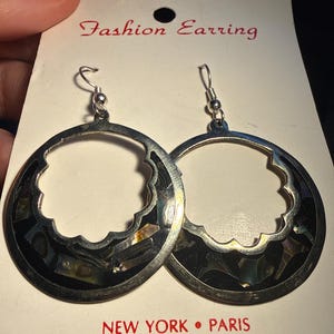 Op de afbeelding: Zilverkleurige oorringen met een geschulpte rand en ingelegde zwarte en iriserende schelpstukken. De oorbellen worden getoond op een witte kaart met de tekst "Fashion Earring" en "NEW YORK &bull; PARIS".